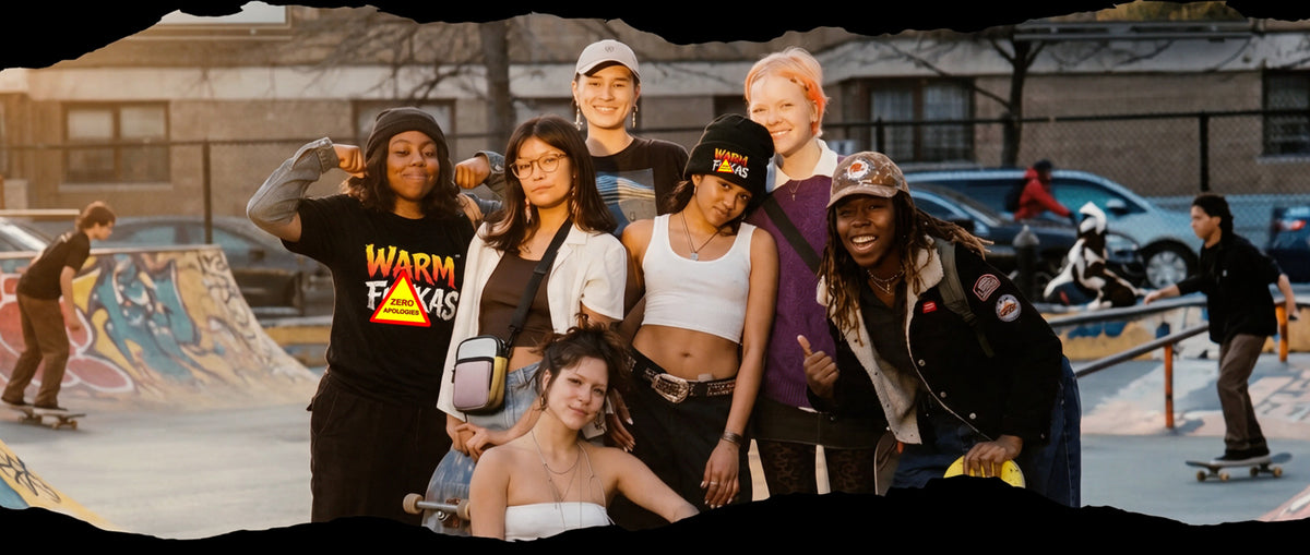 A group of seven young adults posing together at an outdoor skatepark during golden hour, with other skaters and a ramp visible in the background.